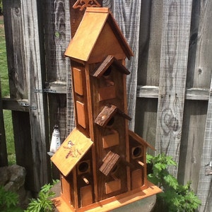 May include: A wooden birdhouse with multiple compartments and a pointed roof. The birdhouse is painted brown and has a natural wood finish. It is mounted on a wooden base.