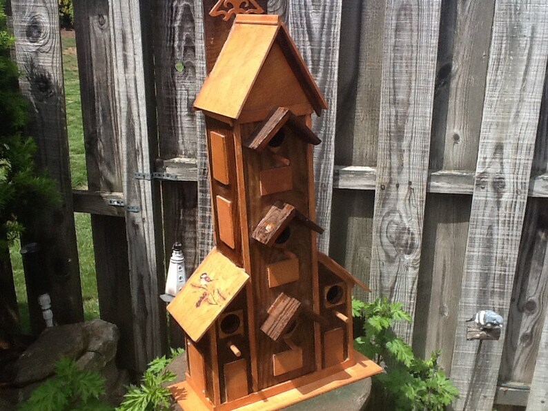 May include: A wooden birdhouse with multiple compartments and a pointed roof. The birdhouse is painted brown and has a natural wood finish. It is mounted on a wooden base.