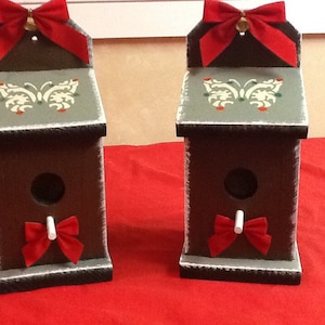 May include: Two brown wooden birdhouses with a gray painted butterfly design on the top. Each birdhouse has a red bow on the front and a white stick protruding from the hole.