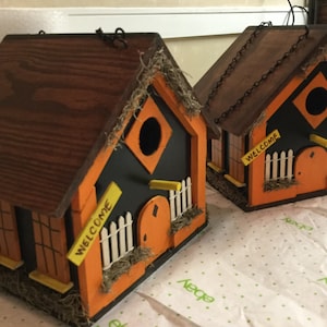 May include: Two wooden birdhouses painted in black, orange, and yellow with a welcome sign on each. The birdhouses have a small white picket fence and a yellow door.
