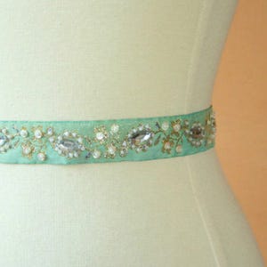 Puede incluir: Una cinta de satén verde claro con hilo dorado y diamantes de imitación transparentes. La cinta está decorada con pequeñas perlas blancas y tiene un diseño floral.