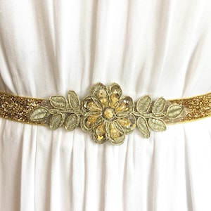 May include: A gold glitter belt with a gold embroidered flower embellishment. The flower has a centre of gold sequins and is surrounded by gold leaves.