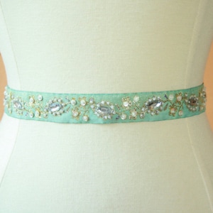 Puede incluir: Una cinta de satén verde claro con adornos dorados y plateados, incluyendo pedrería y perlas. La cinta está decorada con un patrón floral.
