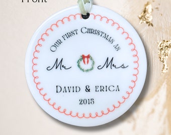 Könnte beinhalten: Runder weißer Keramik-Anhänger mit roten und weißen Nähten am Rand. Der Anhänger trägt die Aufschrift "Our First Christmas as Mr. & Mrs. David & Erica 2015".