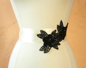 May include: A white satin sash with a black beaded floral embellishment. The embellishment features three large flowers with leaves.