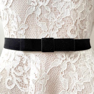 May include: A black velvet belt with a bow detail. The belt is worn over a white lace dress. The bow is centered and rectangular. The lace dress has a floral pattern.