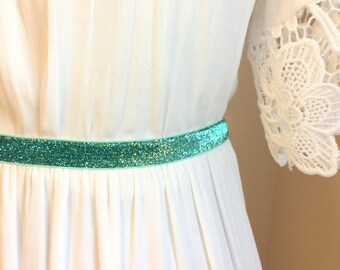 Könnte beinhalten: Ein weißes Kleid mit einem türkisfarbenen Glitzerrband. Das Kleid hat einen Spitzenbesatz an den Ärmeln.
