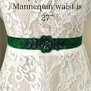 Puede incluir: Una faja de cuentas verdes con un diseño floral y hojas verdes. La faja mide 27 pulgadas de largo y se muestra sobre un vestido de encaje blanco.