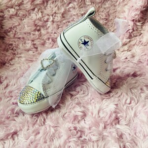 May include: A pair of white baby Converse sneakers with rhinestones and a white satin ribbon. The sneakers have the Converse logo on the side.