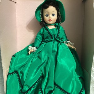May include: A vintage doll wearing a green satin dress with black trim and a green bonnet. The doll has dark hair and blue eyes.