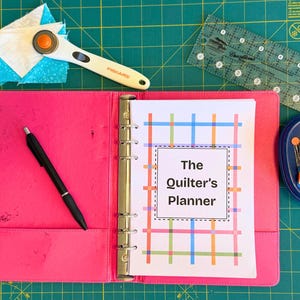 May include: A bright pink binder titled The Quilter's Planner with a colorful grid pattern on the cover page, accompanied by a black pen, a Fiskars rotary cutter, a seam ripper, a clear acrylic ruler, and a blue container filled with sewing pins.
