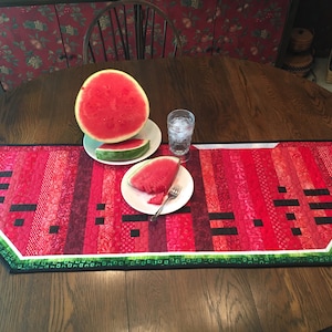 May include: A watermelon-themed table runner with red, green, and black fabric strips. The runner is on a wooden table with a watermelon slice on a white plate.