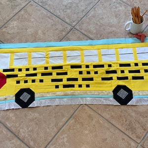 Op de afbeelding: Een gele schoolbusdeken met zwarte geruite ramen, een rood stopteken en zwarte wielen. De deken ligt op een betegelde vloer met twee groene appels op de voorgrond.