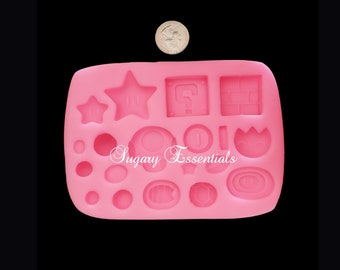 May include: A pink silicone mold featuring various shapes from the Super Mario video game series, including stars, question mark blocks, coins, and mushrooms. The mold has the text "Sugary Essentials" in a cursive font. A quarter is placed above the mold.