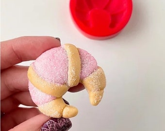 May include: A pink and yellow croissant-shaped silicone mold for baking or crafting. The mold is shown with a pink and yellow fondant croissant.