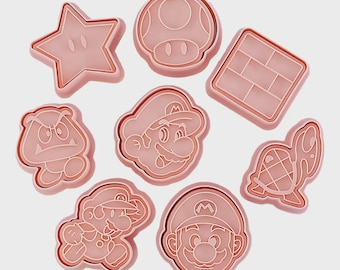May include: A collection of eight pink plastic cookie cutters in various shapes, including a star, mushroom, brick, Goomba, Mario's face, running Mario, and a turtle. These cutters are designed for making cookies or other treats.