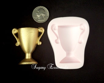 May include: A gold-colored trophy next to a quarter and a pink silicone mold of a trophy. The trophy has two handles and a wide base. The mold is in the shape of the trophy. The background is black.