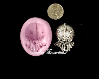 May include: A pink silicone mold for making a small, stitched-face doll. The mold is shaped like a head and has a detailed design. A silver doll is shown next to the mold.