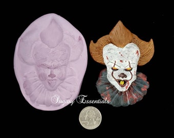May include: A silicone mold for making a cake topper or other edible decoration. The mold is shaped like a clown's face with a red nose, white face paint, and a red and black ruffled collar. The mold is next to a finished cake topper, which is painted in a similar style. The cake topper is about the size of a US quarter.
