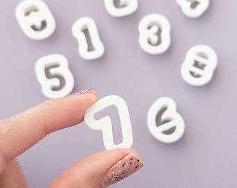 May include: White number-shaped cookie cutters, including the number one being held by a person. The numbers are scattered on a light purple surface. The cookie cutters are made of a solid white material.