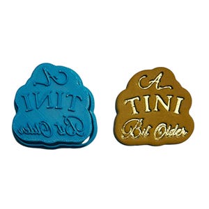Puede incluir: Un cortador de galletas de plástico azul con el texto "A TINI Bit Older" en blanco. Una galleta marrón con el mismo texto en letras doradas.