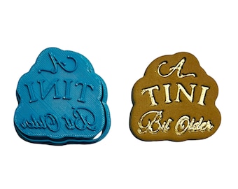 May include: A blue plastic cookie cutter with the text "A TINI Bit Older" in white. A brown cookie with the same text in gold lettering.