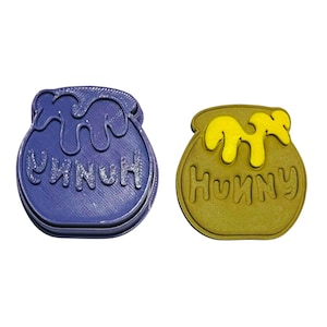 Pode incluir: Dois cortadores de biscoitos em forma de pote de mel do Winnie the Pooh. Um é roxo e diz "Hunny" e o outro é marrom e diz "Hunny".