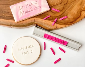 Puede incluir: Una galleta rosa y blanca con el texto "LissleLou AlphaBakes" en ella. Una galleta blanca con el texto "AlphaBakes Font 2" en ella. Un sello para galletas plateado con el texto "AlphaBakes" en él. Varias letras de plástico rosa están esparcidas alrededor de las galletas.