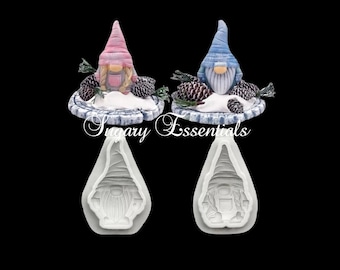 May include: Two silicone molds for making gnome cake toppers. The molds are white and feature a gnome with a pink hat and a gnome with a blue hat. Each gnome is sitting on a snowy base with pine cones.
