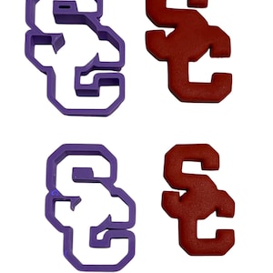 Puede incluir: Dos cortadores de galletas y dos galletas con la forma de las letras "SC". Un cortador es morado, el otro es una galleta roja. Las galletas también son rojas. Las letras son de estilo bloque.