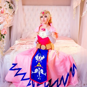 Puede incluir: Una mujer vestida como la princesa Zelda de la serie de videojuegos The Legend of Zelda. Lleva un vestido rosa con una capa azul y dorada, un cintur&oacute;n dorado y una corona dorada. Est&aacute; sentada en una cama con s&aacute;banas blancas y un dosel blanco.