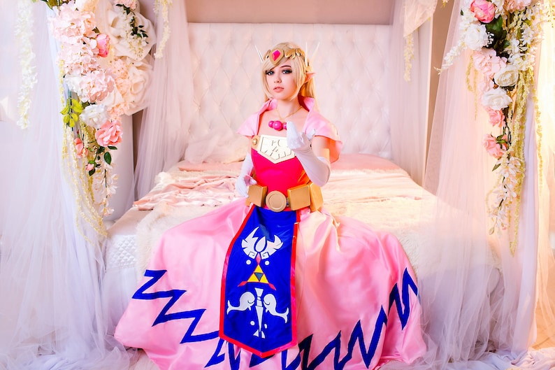 Puede incluir: Una mujer vestida como la princesa Zelda de la serie de videojuegos The Legend of Zelda. Lleva un vestido rosa con una capa azul y dorada, un cintur&oacute;n dorado y una corona dorada. Est&aacute; sentada en una cama con s&aacute;banas blancas y un dosel blanco.