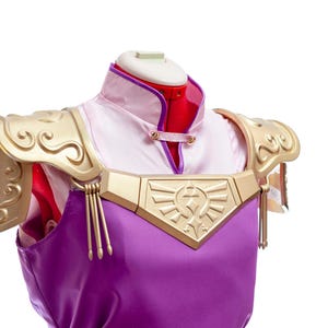 Puede incluir: Un disfraz de cosplay con una t&uacute;nica morada con detalles dorados y hombreras. El disfraz incluye una camisa blanca y rosa con cuello y un emblema dorado en el pecho. Las hombreras tienen un dise&ntilde;o decorativo en espiral.