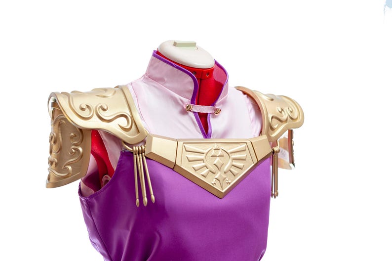 Puede incluir: Un disfraz de cosplay con una t&uacute;nica morada con detalles dorados y hombreras. El disfraz incluye una camisa blanca y rosa con cuello y un emblema dorado en el pecho. Las hombreras tienen un dise&ntilde;o decorativo en espiral.