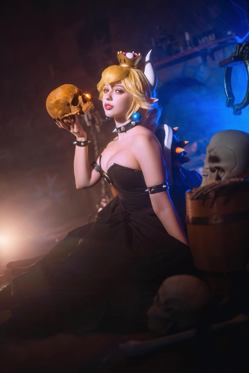 Puede incluir: Una mujer con un vestido negro y una corona, sosteniendo una calavera. Tiene el pelo rubio y pendientes azules. La imagen tiene una est&eacute;tica oscura y teatral, con un accesorio de calavera y un esquema de iluminaci&oacute;n azul y naranja.