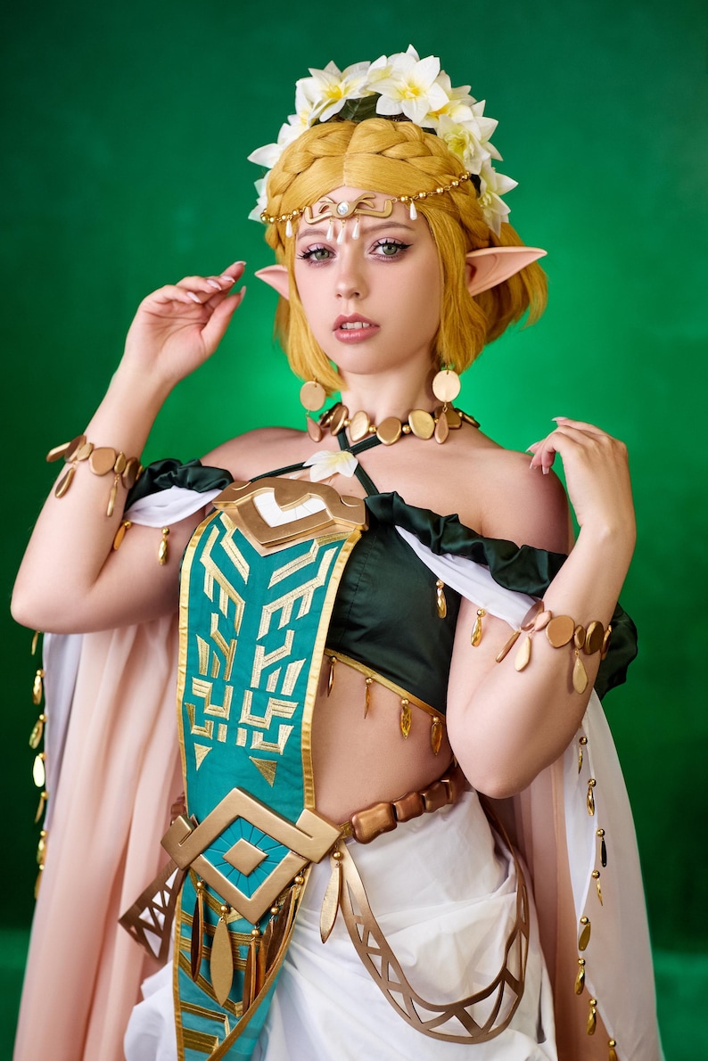 May include: A woman in a cosplay costume with a green and gold bodice, white skirt, and a floral crown. The costume features gold accents, including jewelry and decorative elements. The background is a solid green.