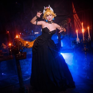 Puede incluir: Una persona con un vestido negro sin tirantes y una corona, con cabello rubio y pendientes azules, posa en una habitaci&oacute;n con poca luz y velas. El vestido tiene un corpi&ntilde;o ajustado y una falda fluida. El tema general es el cosplay.