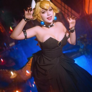 Puede incluir: Una mujer con un vestido negro y una peluca rubia, vestida como un personaje de videojuego. Lleva una corona, un collar de p&uacute;as y pulseras. El vestido tiene un escote coraz&oacute;n y un detalle de anillo dorado. Pendientes azules y esmalte de u&ntilde;as negro completan el look.