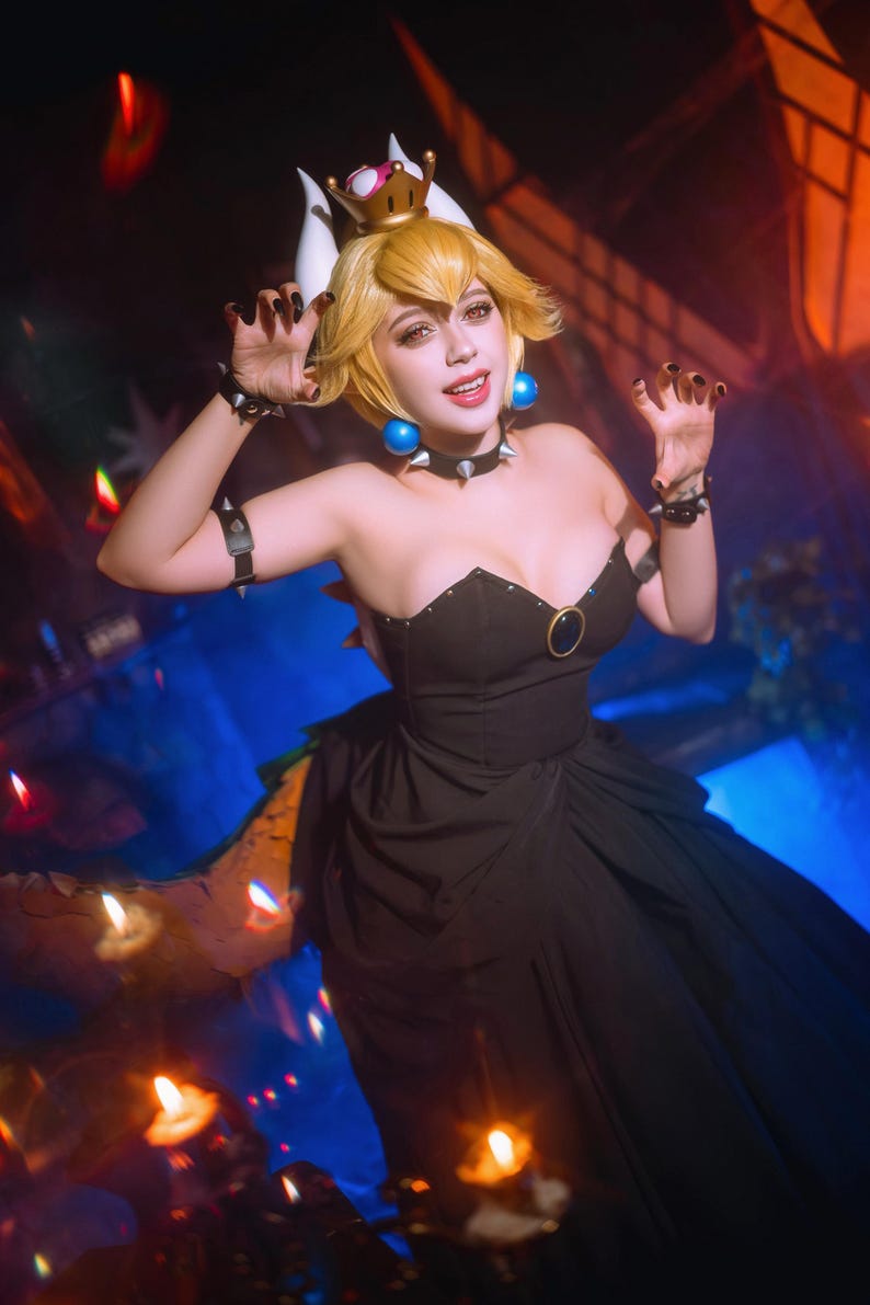 Puede incluir: Una mujer con un vestido negro y una peluca rubia, vestida como un personaje de videojuego. Lleva una corona, un collar de p&uacute;as y pulseras. El vestido tiene un escote coraz&oacute;n y un detalle de anillo dorado. Pendientes azules y esmalte de u&ntilde;as negro completan el look.