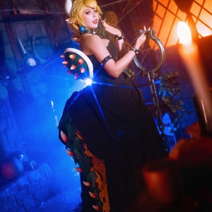 Puede incluir: Una mujer con un vestido negro con un accesorio en forma de caparaz&oacute;n con p&uacute;as, una corona y una cadena. El disfraz est&aacute; iluminado con luces azules y naranjas, creando un efecto dram&aacute;tico. El tema general es el cosplay.