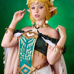 May include: A woman in a cosplay costume with a green and gold bodice, white skirt, and a floral crown. The costume features gold accents, including jewelry and decorative elements. The background is a solid green.