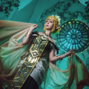May include: A person in a cosplay costume with a gold headpiece, green and gold accents, and flowing fabric. The costume has a geometric design on the bodice. The background is a teal color with floral accents and a stained glass window.