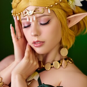 May include: A person with blonde hair styled in a braid, wearing a gold headpiece with pearl accents and a matching necklace. The person is also wearing a green top and has elf-like ears. White flowers are in the hair.