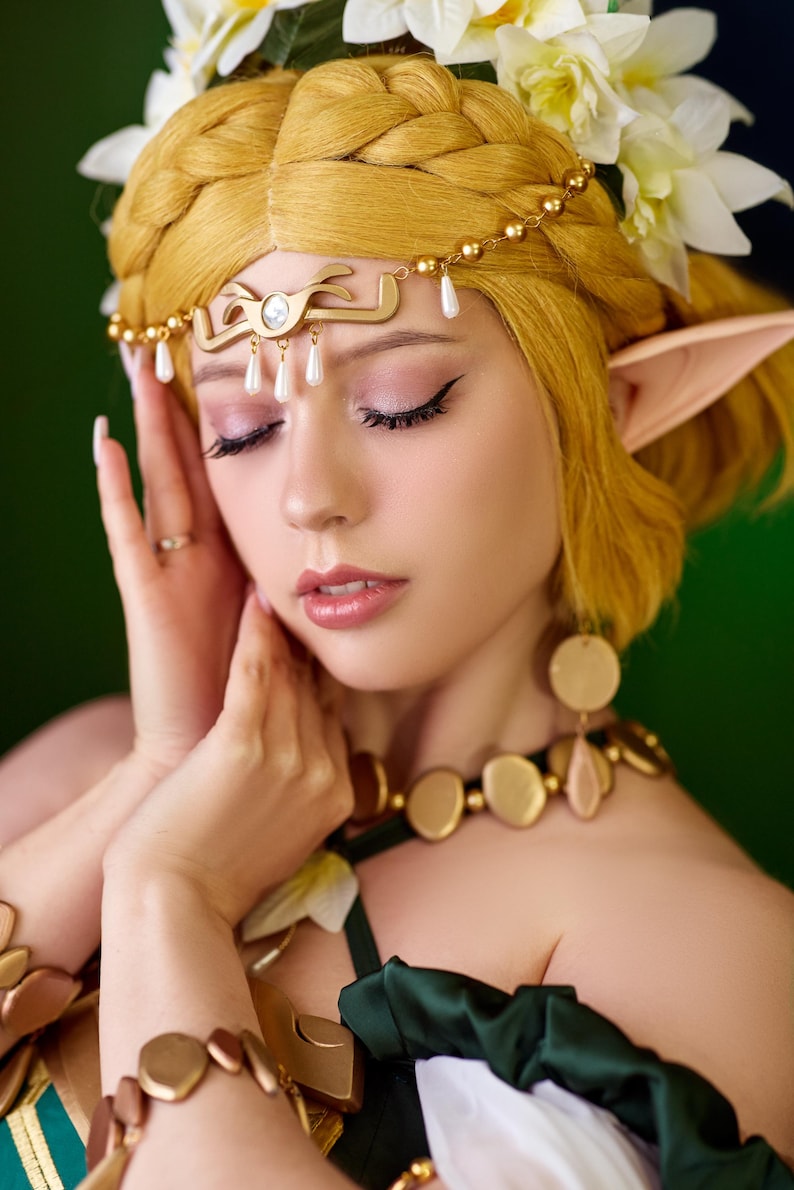 May include: A person with blonde hair styled in a braid, wearing a gold headpiece with pearl accents and a matching necklace. The person is also wearing a green top and has elf-like ears. White flowers are in the hair.