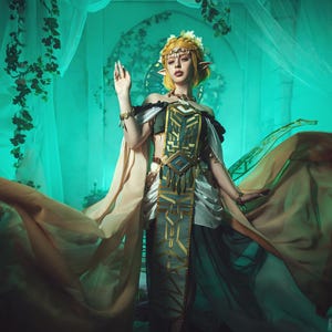May include: A woman in a fantasy costume with long, flowing fabric. The costume features gold, green, and white accents, with a detailed bodice and headpiece. The background is a teal color with vines and sheer curtains.