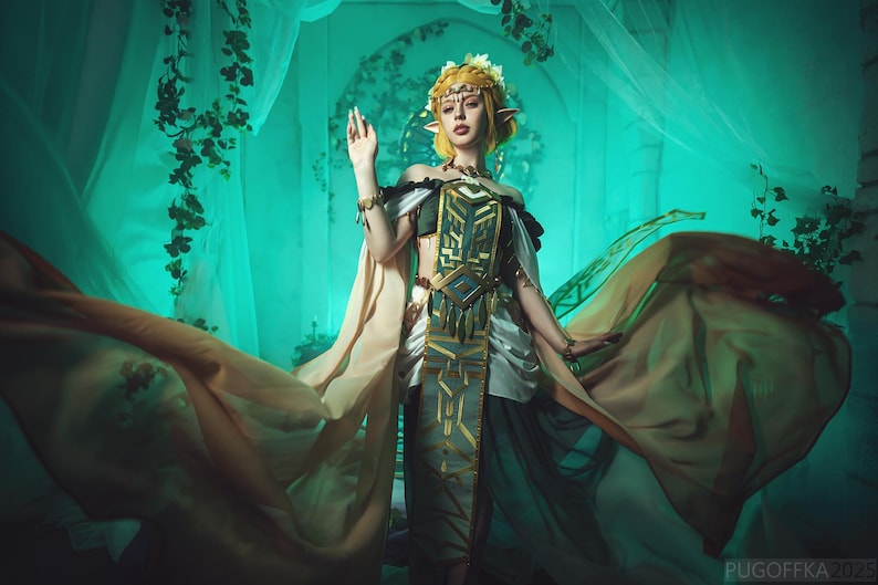 May include: A woman in a fantasy costume with long, flowing fabric. The costume features gold, green, and white accents, with a detailed bodice and headpiece. The background is a teal color with vines and sheer curtains.
