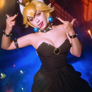 Puede incluir: Un atuendo de cosplay con un vestido negro con escote coraz&oacute;n y un acento dorado. El disfraz incluye una corona, gargantilla y pulseras con p&uacute;as. El personaje tiene el pelo rubio y hace un gesto con la mano.