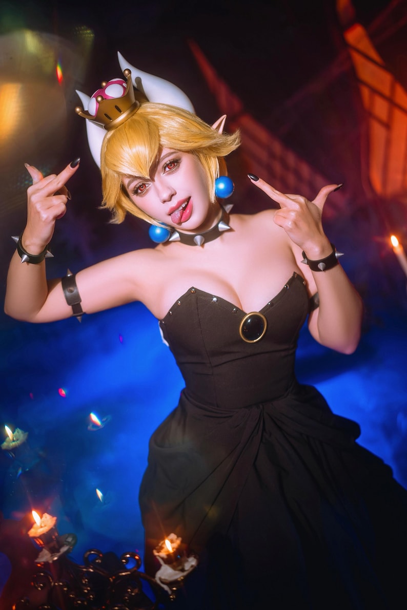 Puede incluir: Un atuendo de cosplay con un vestido negro con escote coraz&oacute;n y un acento dorado. El disfraz incluye una corona, gargantilla y pulseras con p&uacute;as. El personaje tiene el pelo rubio y hace un gesto con la mano.