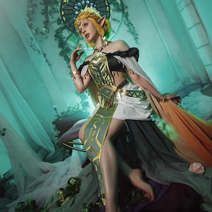 May include: A woman in an elaborate fantasy costume with blonde hair and pointed ears. The dress is a mix of black, white, gray, and gold, with a flowing train in shades of orange and gray. The background is a teal-colored room with floral accents.