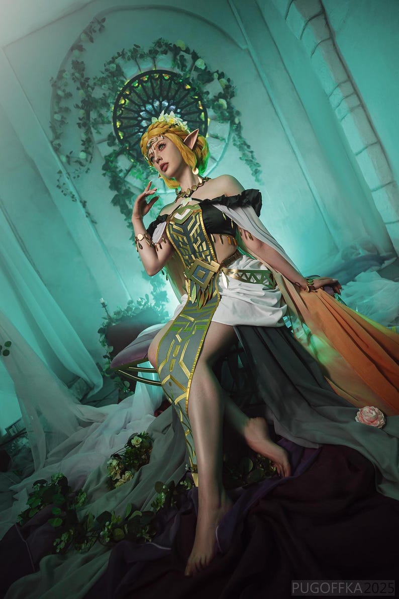 May include: A woman in an elaborate fantasy costume with blonde hair and pointed ears. The dress is a mix of black, white, gray, and gold, with a flowing train in shades of orange and gray. The background is a teal-colored room with floral accents.